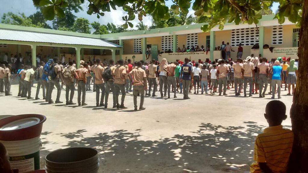 Dans la cour de l'école, de très nombreux enfants sont alignés pour faire l'appel, ils sont face aux salles de classe